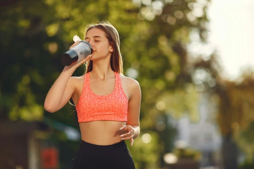 Mulher em roupa fitness bebendo água ao ar livre após o treino, representando a importância da hidratação no treino.